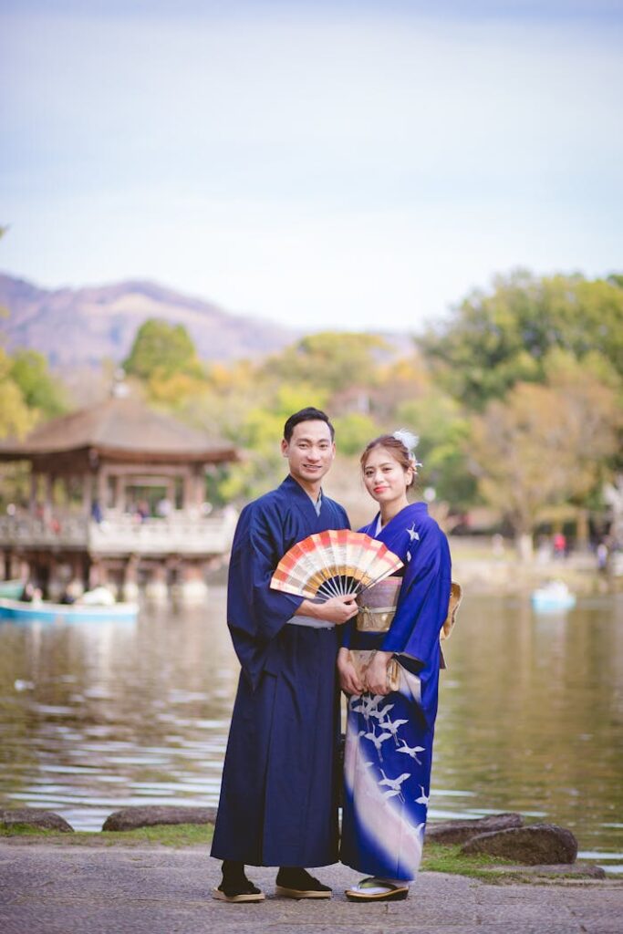 A couple wearing traditional Japanese kimonos stands by a scenic lake, holding a colorful fan.